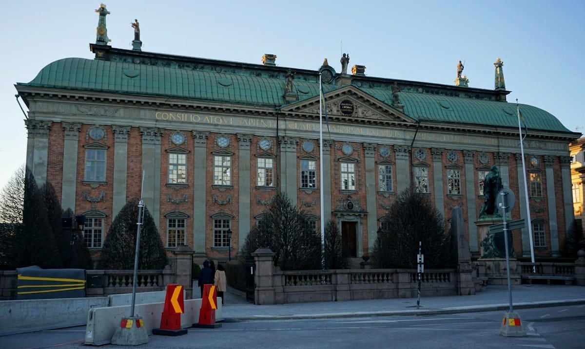 Historic stone building with green roof and columns, lit at dusk with cones and barriers in front