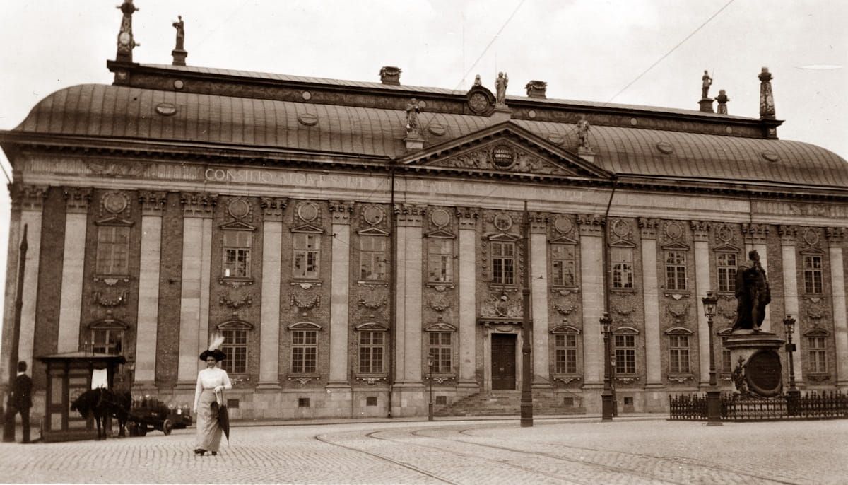 Sepia photo of a grand neoclassical building with columns and a statue in front, people walking nearby