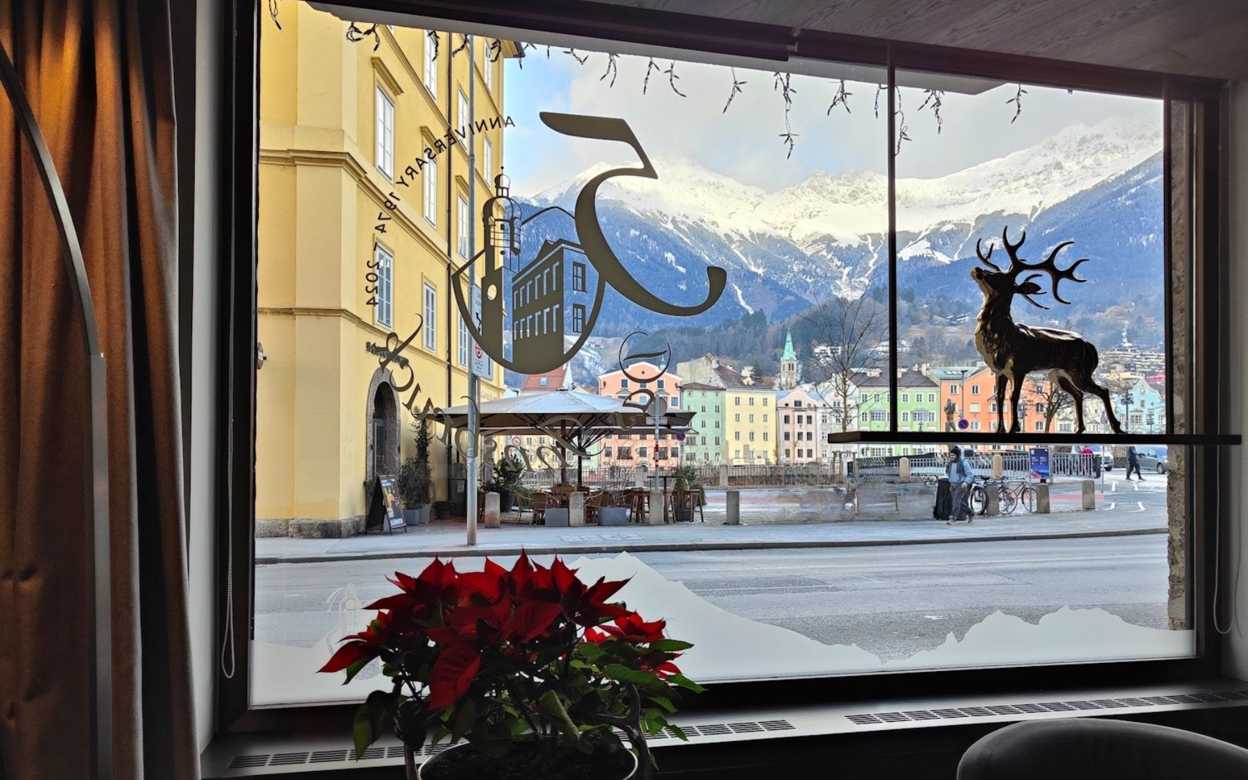 Window view: snow-covered street and mountains, a deer silhouette on the glass, red flowers in foreground, buildings and people outside.