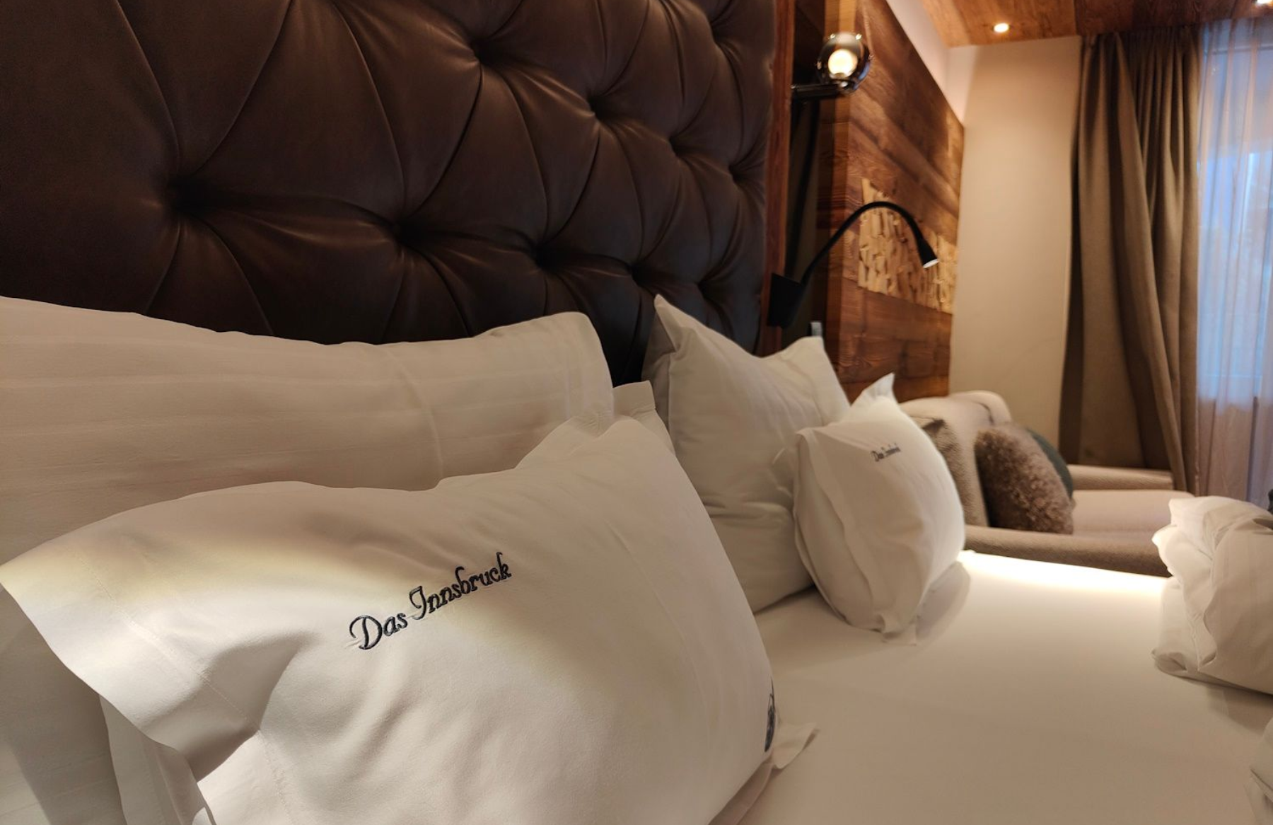 Cozy hotel bed with white linens, pillows, and a tufted leather headboard. Warm lighting and wood accents.