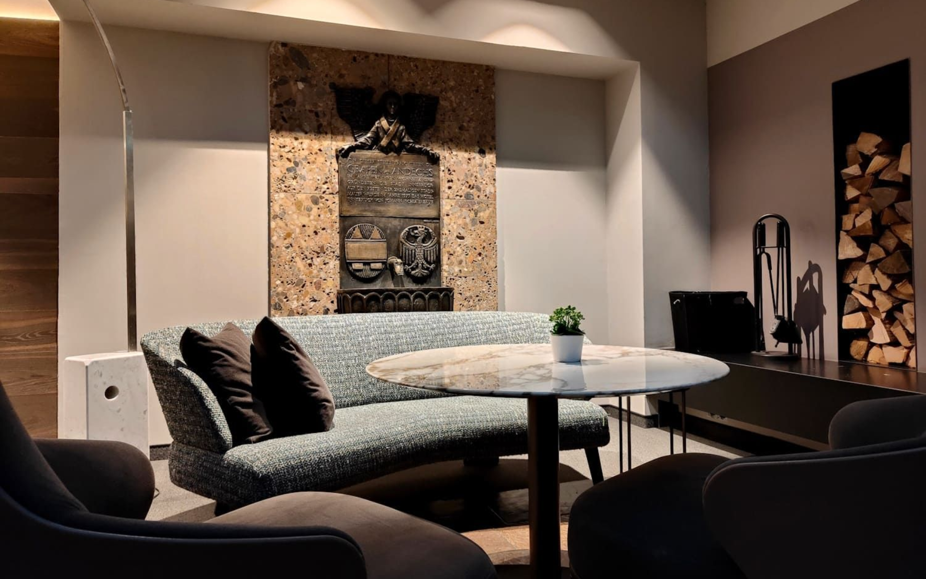 Cozy lounge area with curved blue sofa, round table, and decorative stone artwork. Firewood storage and a small potted plant are also present.