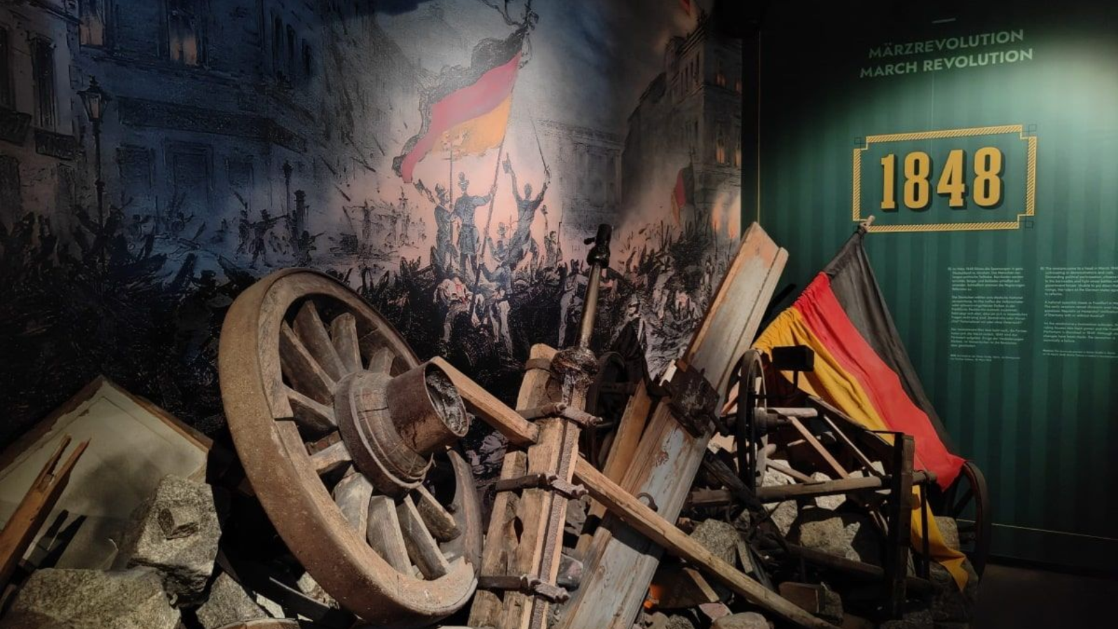 Deutschland Museum Berlin Exhibit about 1848, with a wheel, wooden structures, a German flag, and a mural in a dark setting.