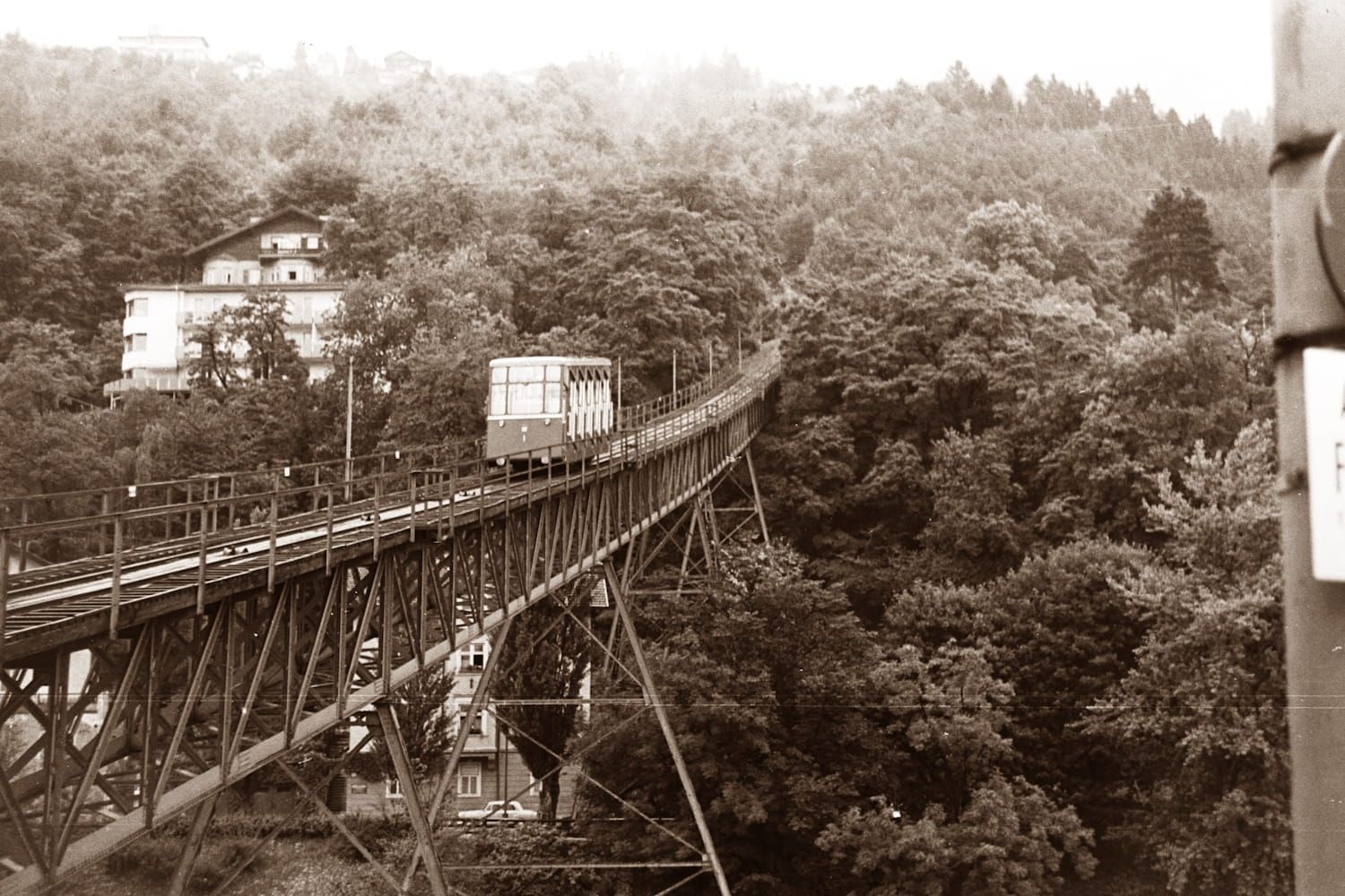 Cable car traversing a bridge, surrounded by trees. A building is visible on the left.