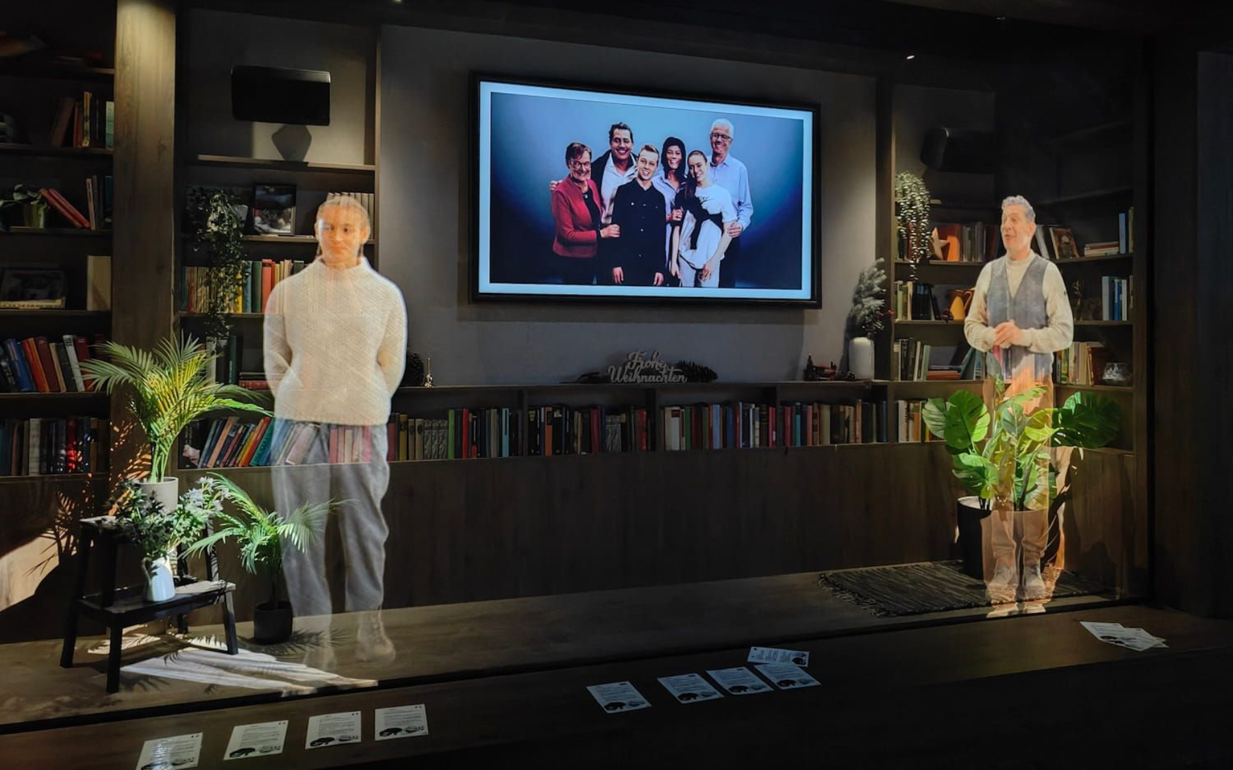Two figures stand on a stage, with a large screen displaying a family photo. Dark room with bookshelves.