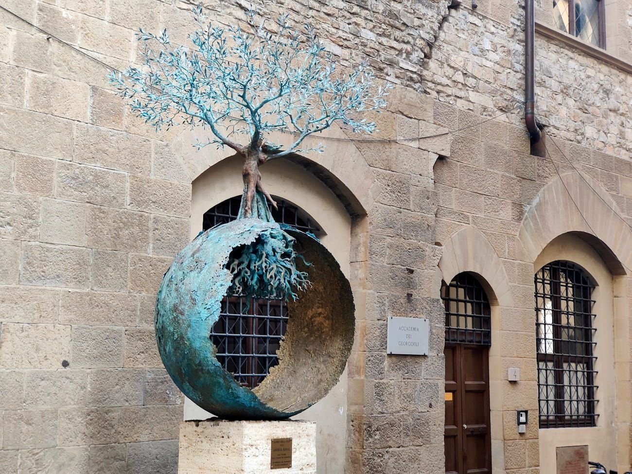 A bronze sculpture of a tree growing out of a hollow sphere, set against a stone building with arched doorways.