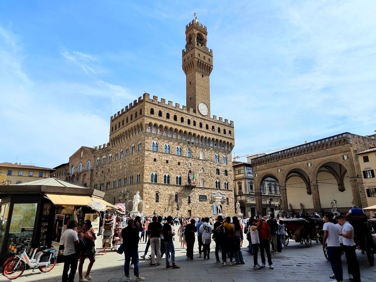 The Palazzo Vecchio rises over the bustling Piazza della Signoria in Florence, with tourists gathered in the square.