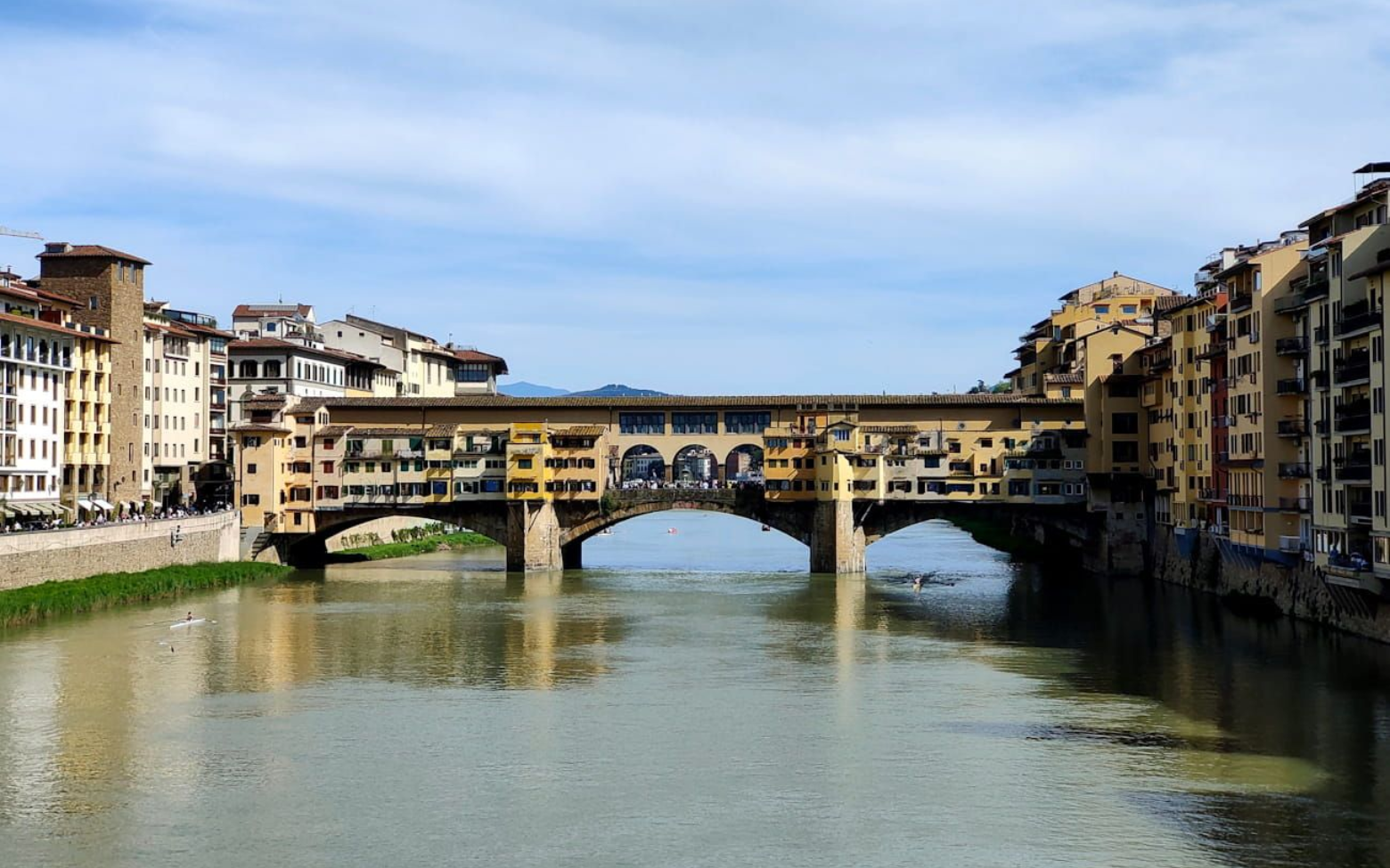 The Ponte Vecchio stone arch bridge with shops spanning the Arno River in Florence, Italy, under a blue sky.