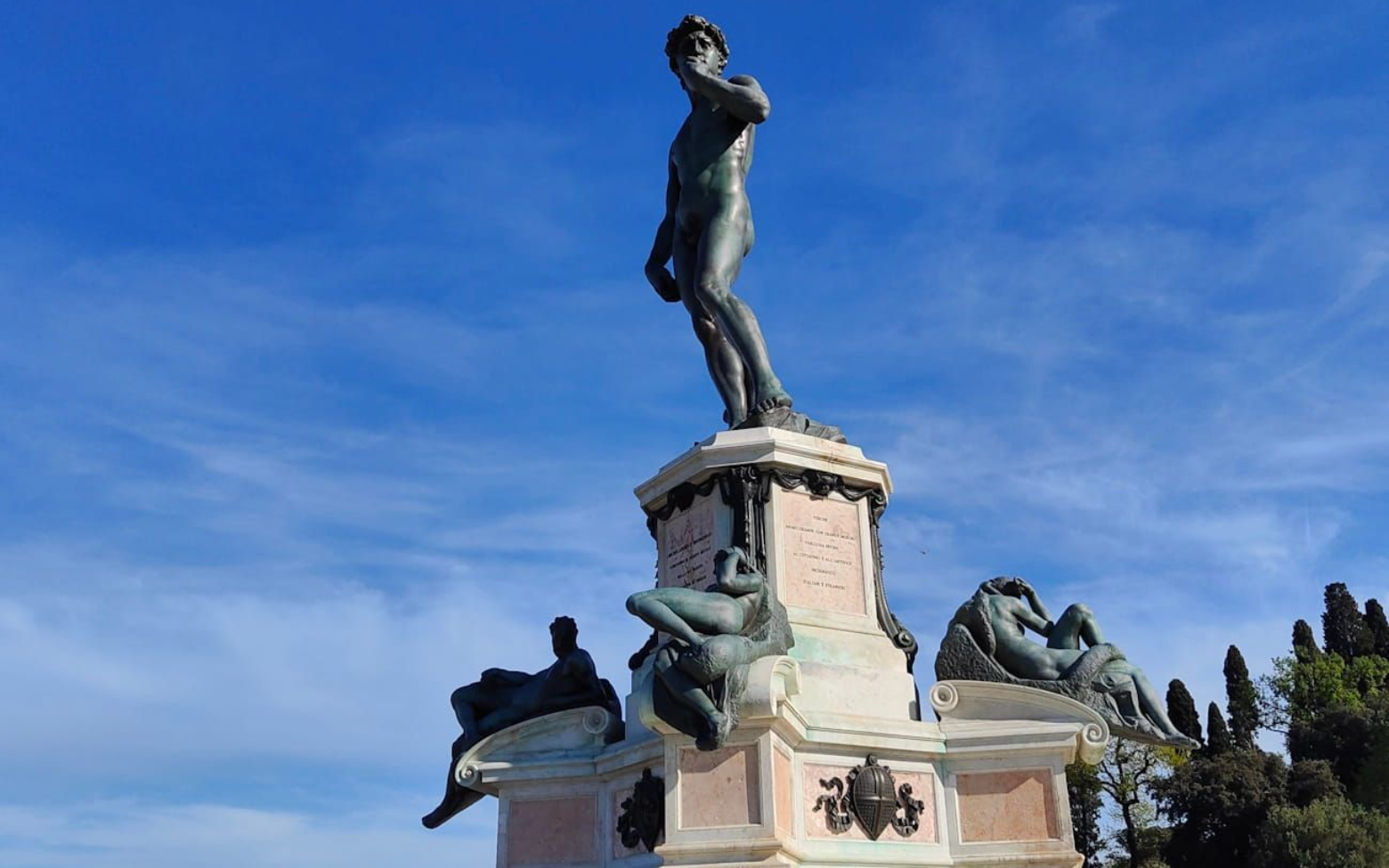 Bronze statue of David atop a stone pedestal with reclining figures, set against a bright blue sky.