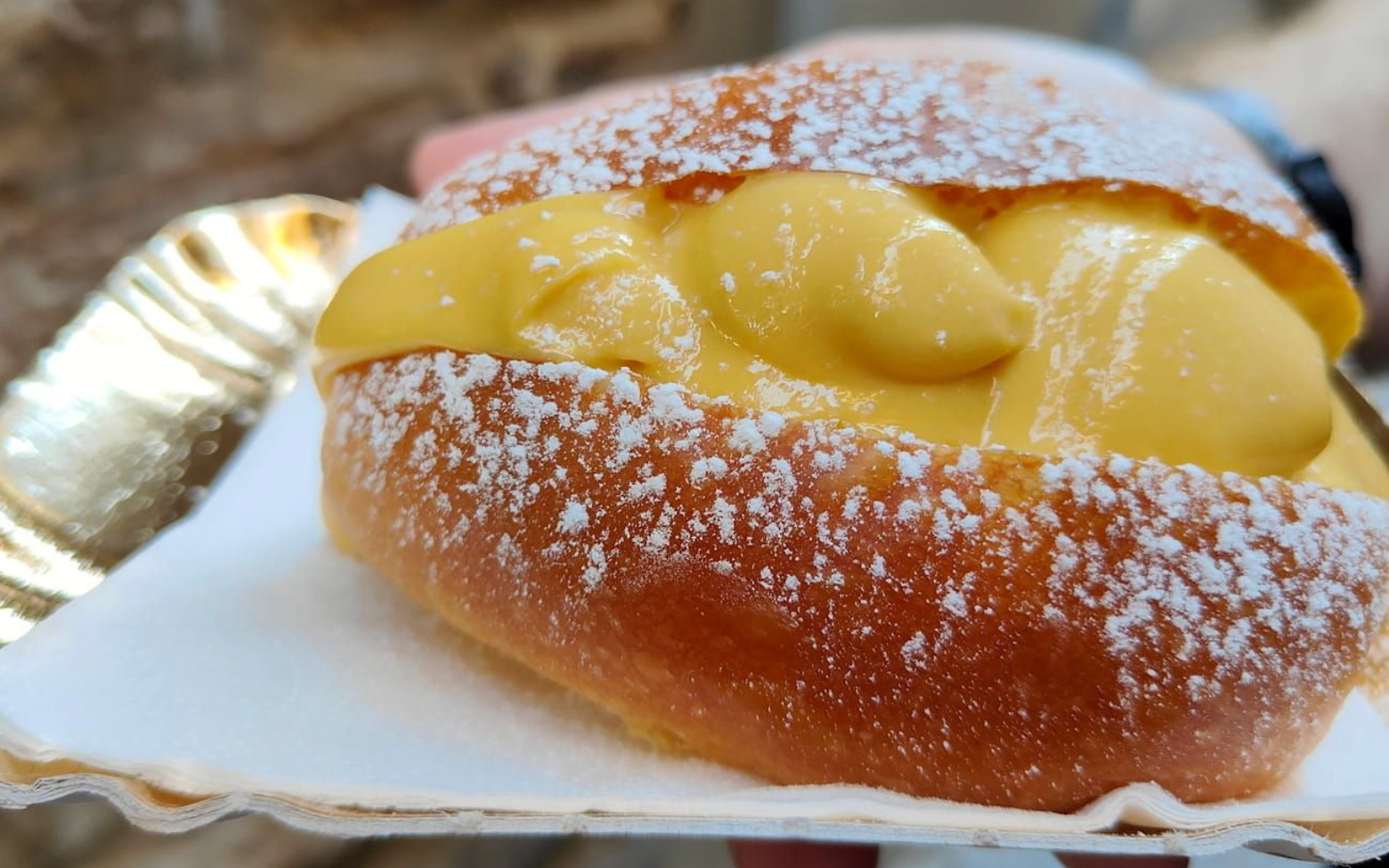 A soft, golden-brown brioche bun filled with smooth yellow cream and dusted with powdered sugar on a paper tray.