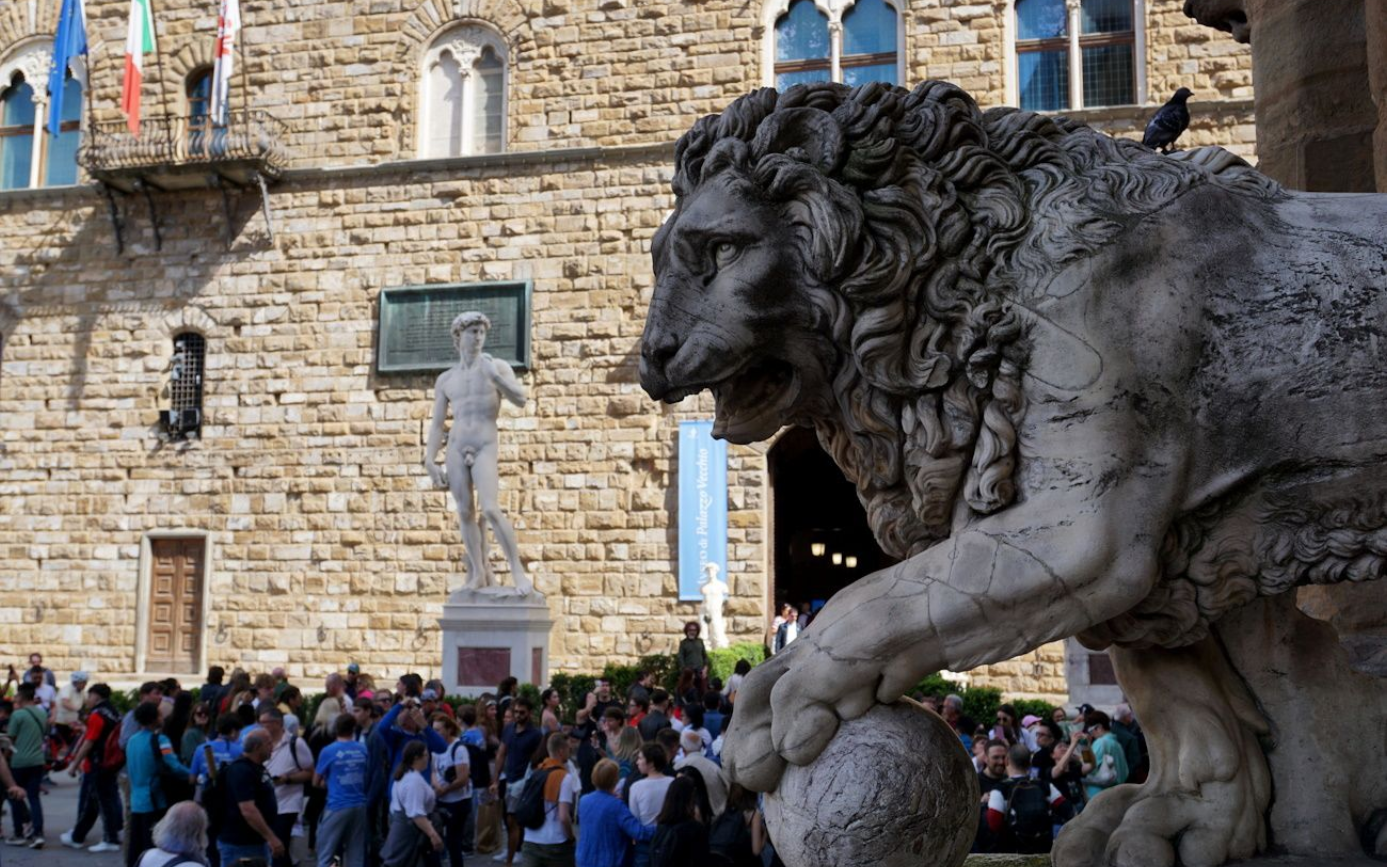 A crowd gathers in front of the Palazzo Vecchio in Florence, featuring a marble lion sculpture and David statue.