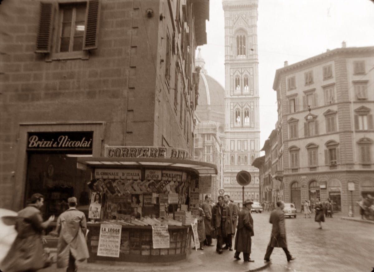 A sepia-toned street scene in Florence featuring a newsstand, pedestrians in coats, and Giotto's Campanile in the distance.