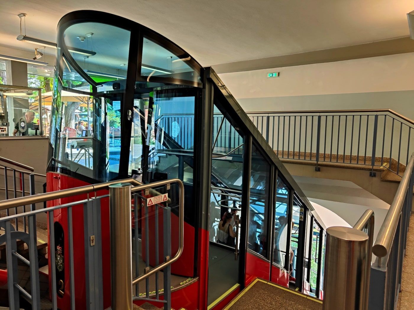 A red cable car ascending a ramp inside a building, with glass walls and a view of the outside. Stairs and railings are visible.
