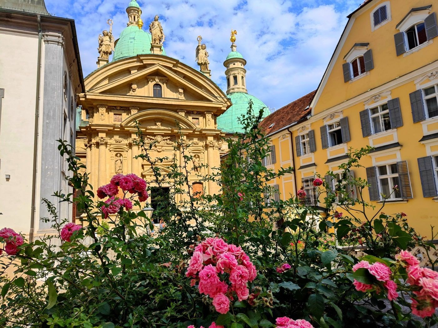 Pink roses in front of an ornate yellow building with green domes and a yellow building to the right under a blue sky.