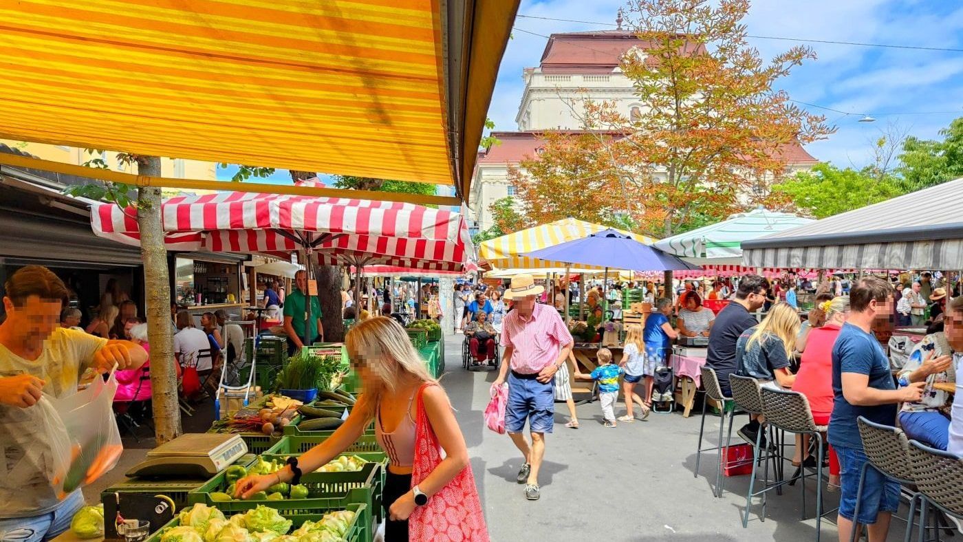 A bustling outdoor market with people shopping at stalls selling produce under colorful awnings.