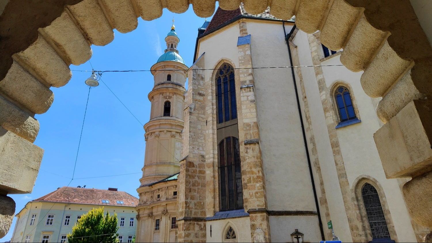 View through an arched doorway of a tall church with a green-topped tower against a blue sky.