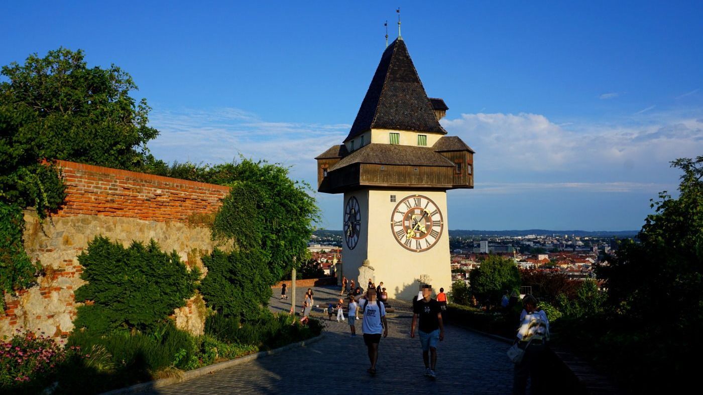 Clock tower in Graz, Austria, with a prominent clock face, overlooking the city on a sunny day. People walk on the pathway.