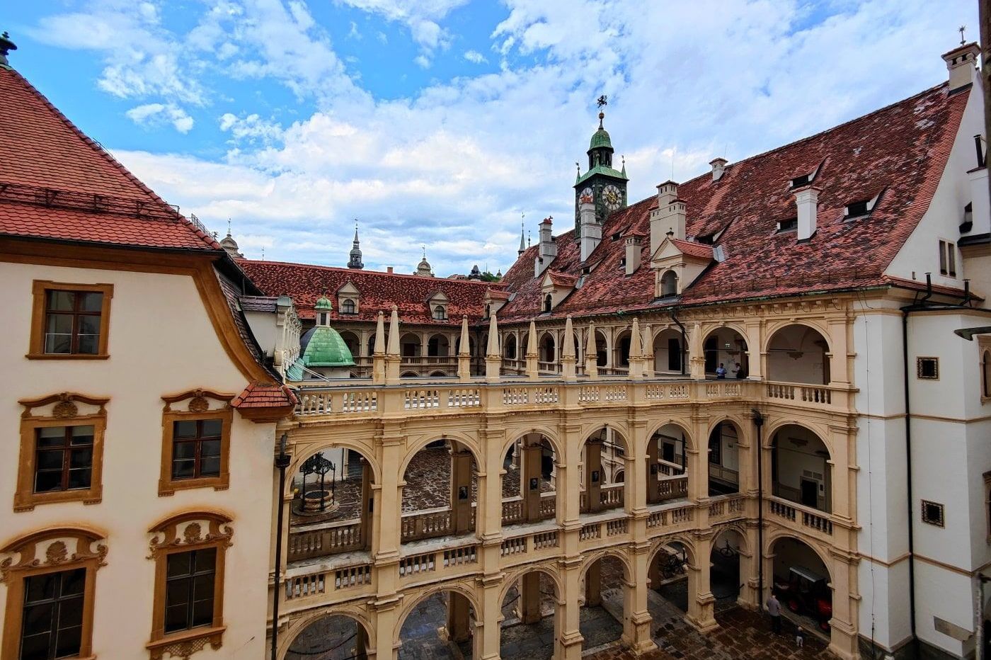 Courtyard of Eggenberg Palace in Graz, Austria, with arched walkways, red-tiled roofs, and a blue sky with scattered clouds.
