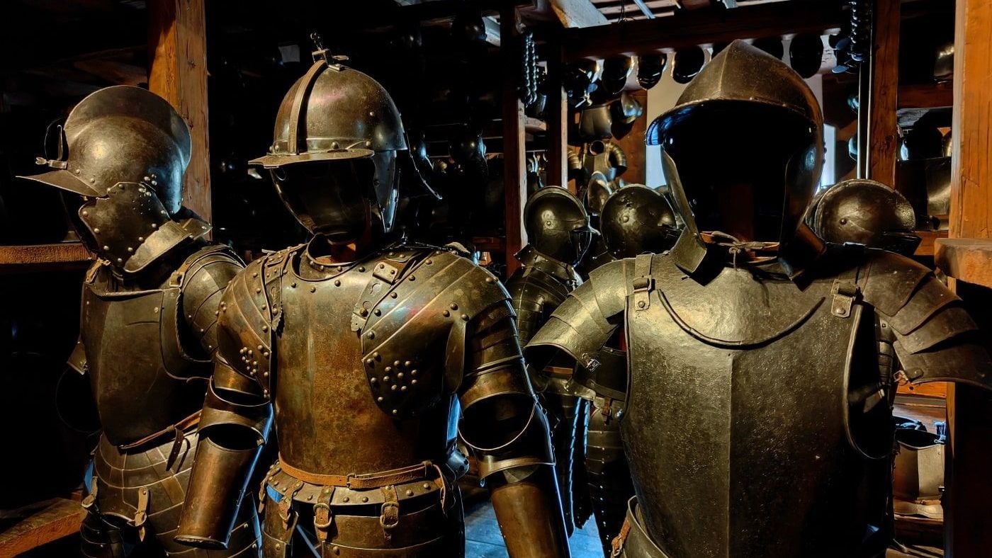 Several suits of armor in a dimly lit room, likely a museum or storage space. They are dark metal, displayed upright.