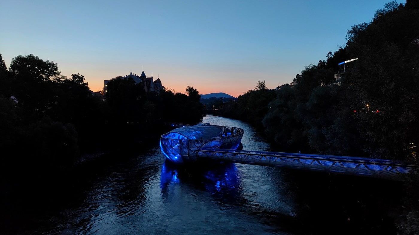A river with a blue-lit structure flows through a dark forest at dusk. Distant buildings are silhouetted in the orange and blue sky.