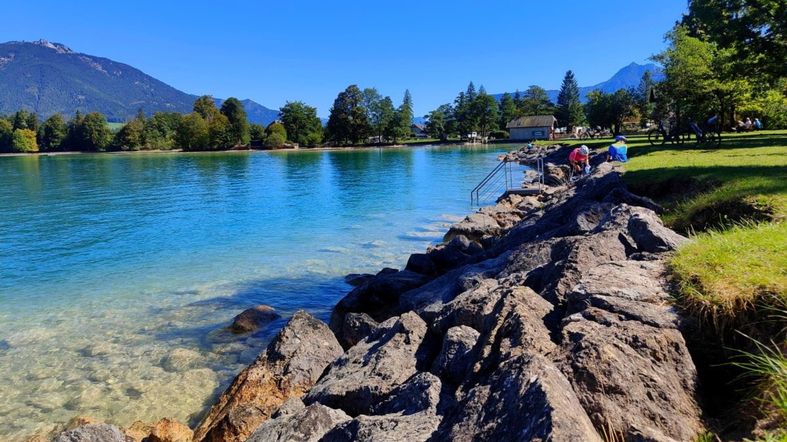 Rocky shoreline with turquoise water, mountains, and a clear blue sky.