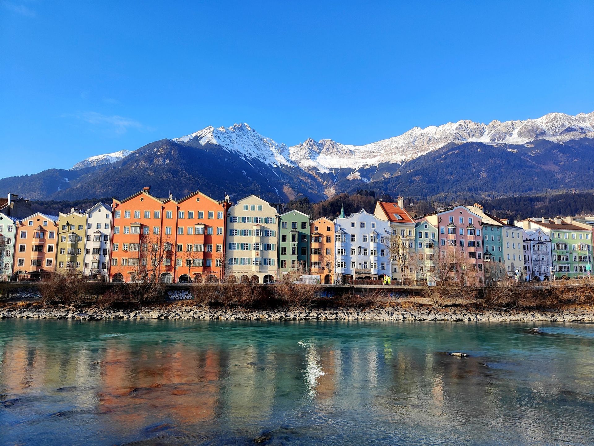 Colorful buildings line a river with snow-capped mountains in the background under a blue sky.