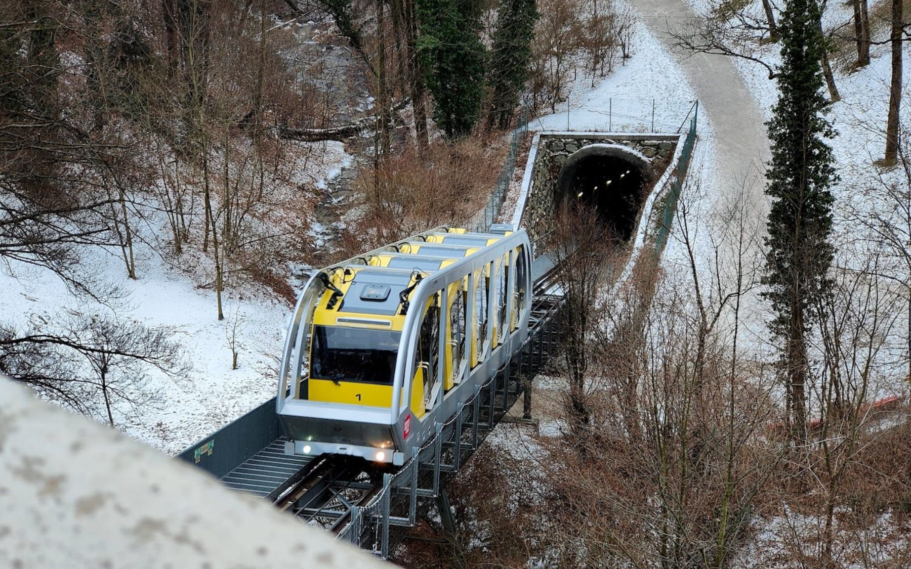 Yellow funicular train on tracks near snow-covered trees and a tunnel entrance.