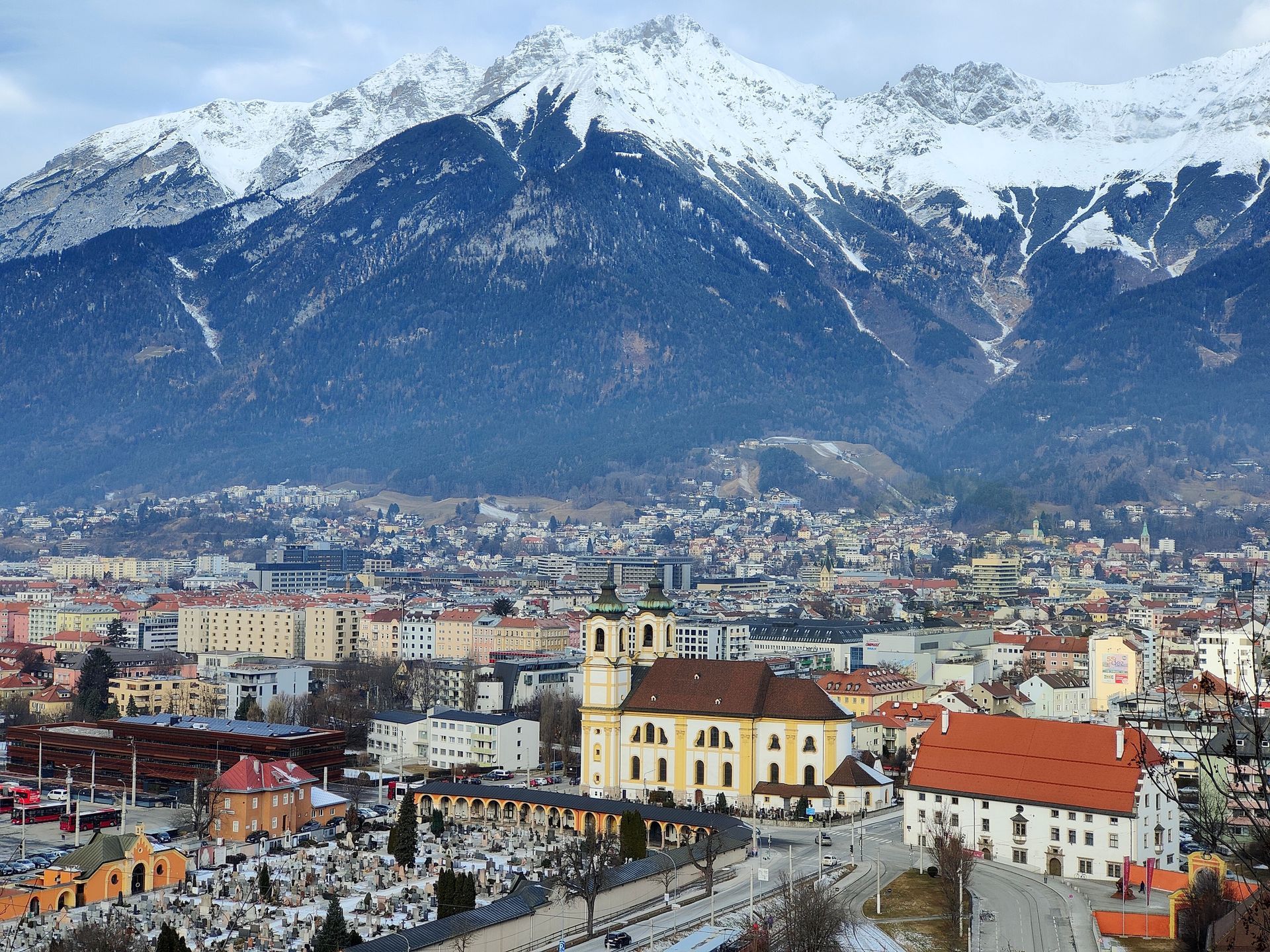 City nestled in valley, snow-capped mountains in background. Yellow church and buildings prominent; overcast sky.