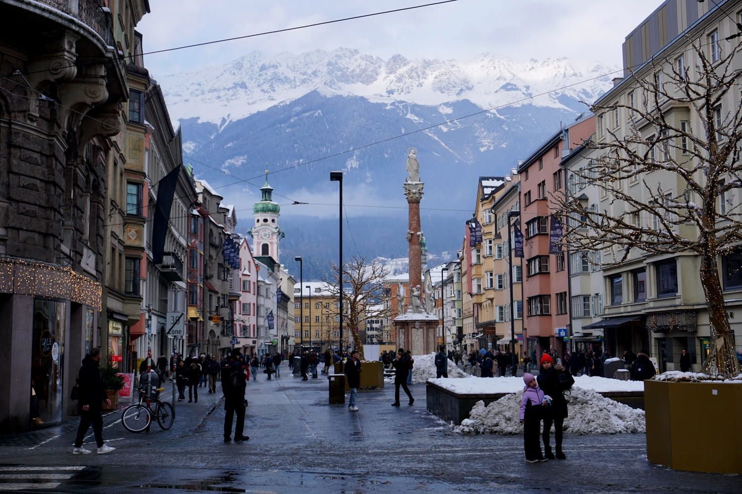 City street scene, snowy mountains in background. Buildings, pedestrians, and a monument.