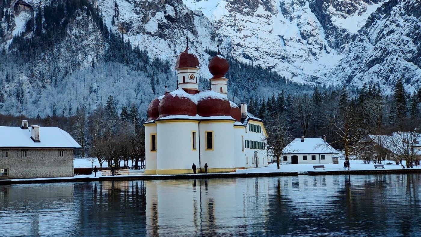 A small church sits in the middle of a lake surrounded by snow covered mountains