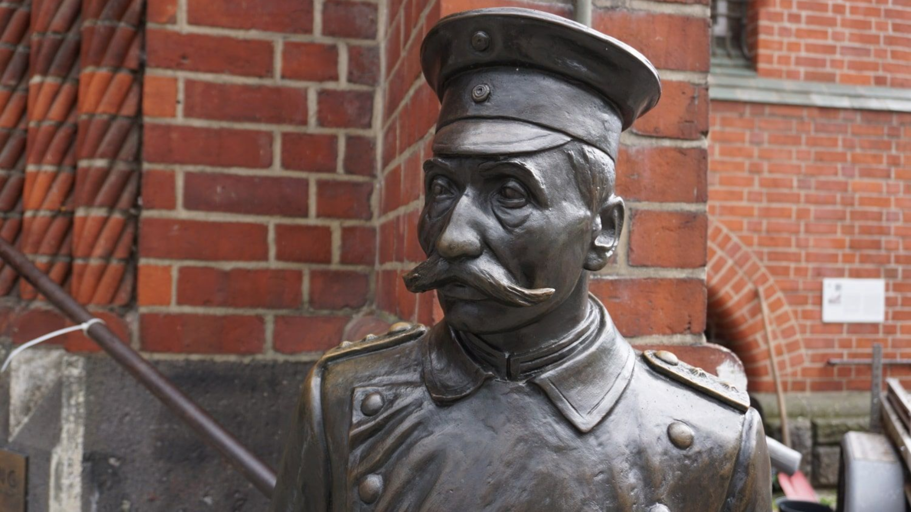 Berlin Köpenick Hauptmann Bronze bust of a mustached man in a military uniform and cap, in front of a brick building.