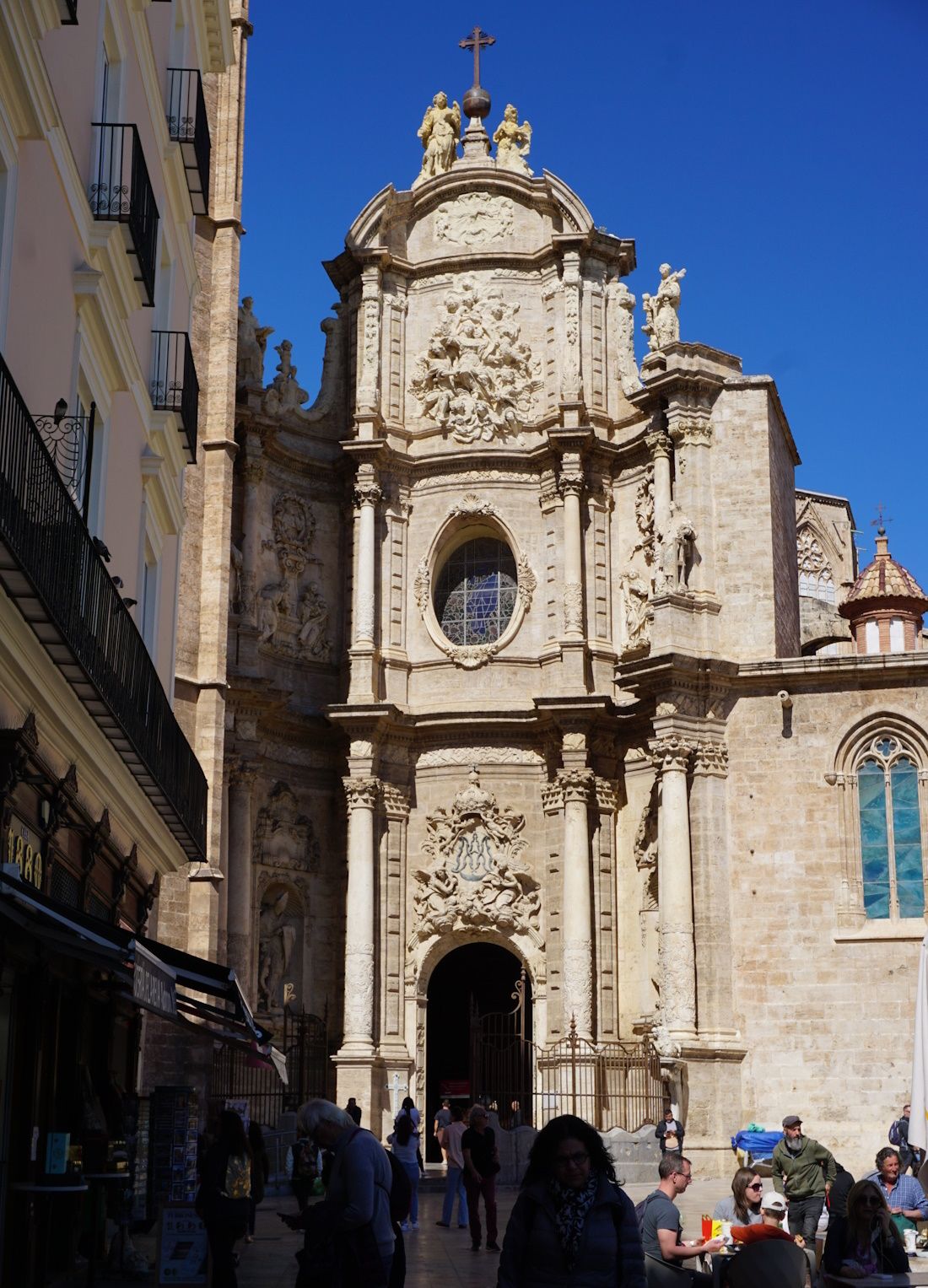 Cathedral facade, beige stone, ornate carvings, arched doorway, pedestrians, outdoor seating, blue sky.