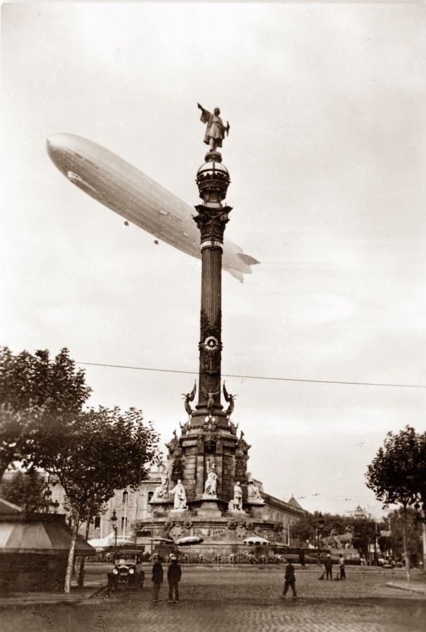 A black and white photo of an airship flying over a statue