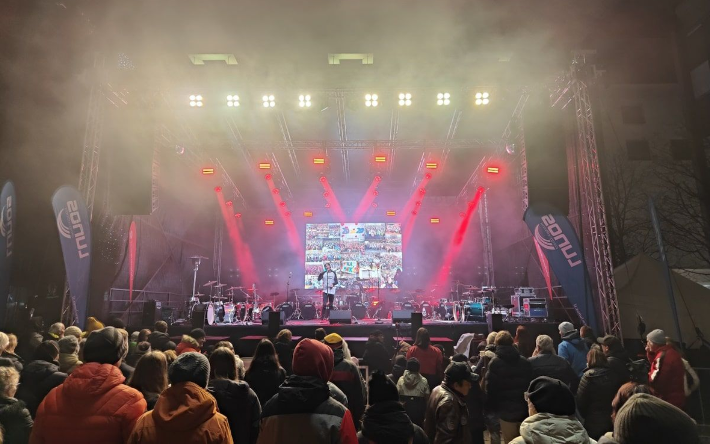 Maribor Konzert Concert stage with performers and crowd, lit with red and white lights. People in winter clothing watch.
