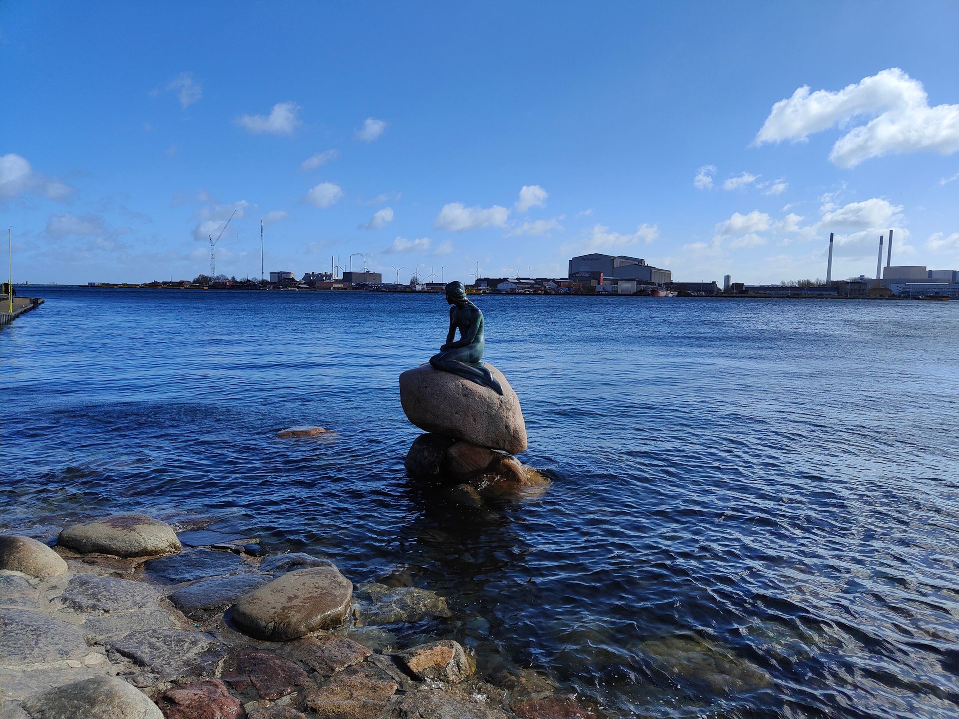 Ein Steinhaufen mit einer Statue an der Spitze im blauen Wasser unter strahlend blauem Himmel, im Hintergrund die ferne Küstenlinie.