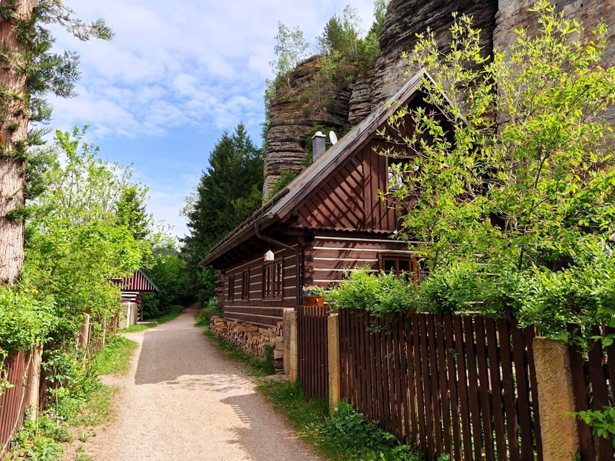 Narrow gravel path beside rustic wooden cottages and lush green trees under a bright blue sky.