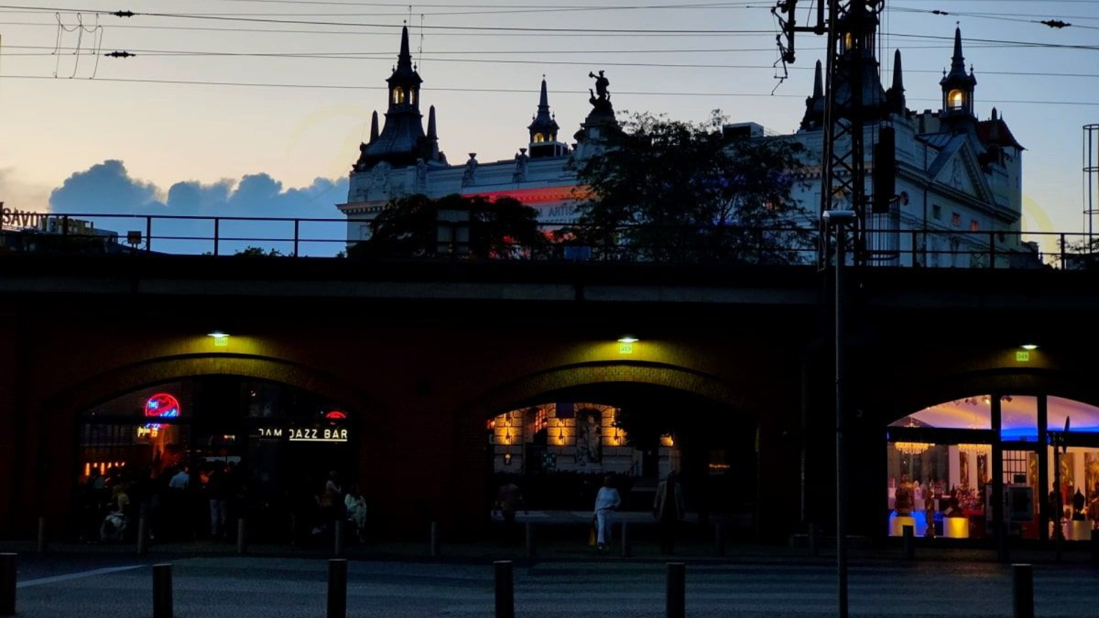 Berlin Night scene of an arched brick bridge with lights, a building with towers in the background.