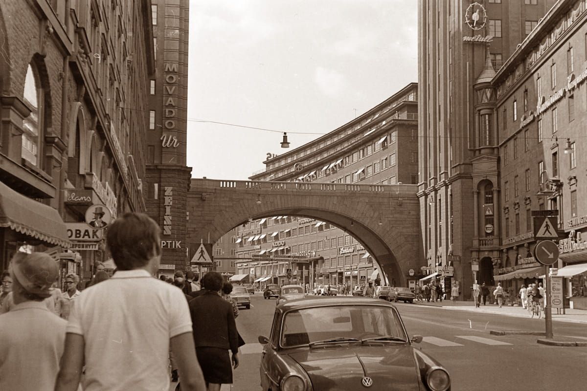 Sepia-toned city street with pedestrians, cars, and an arched bridge between tall buildings