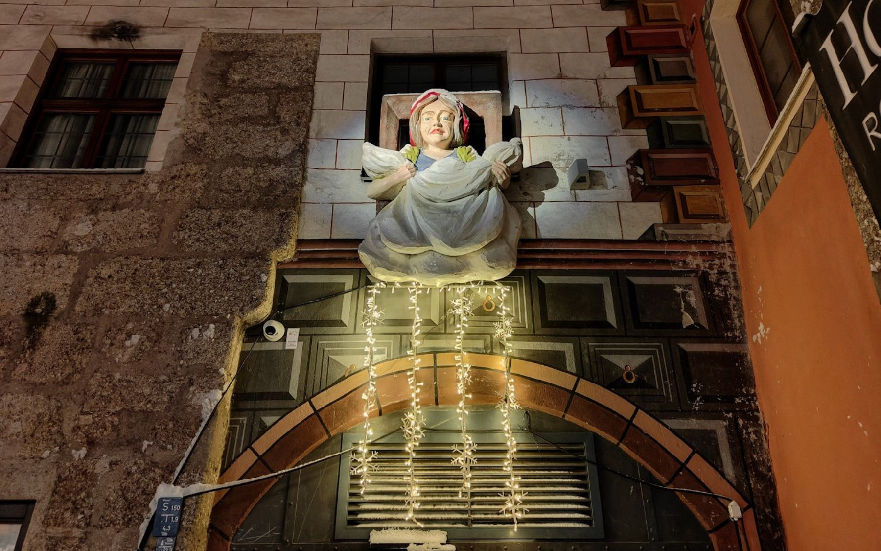 Statue of angel above an arched doorway, lit with fairy lights, on an old building at night.
