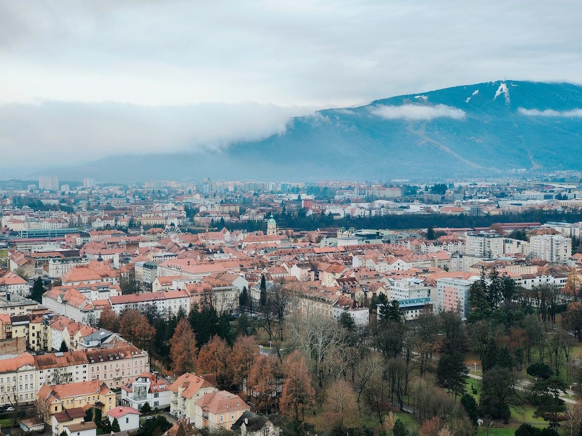 Maribor Aussicht Cityscape with red-tiled roofs, trees, and a mountain with low clouds in the background on a cloudy day.