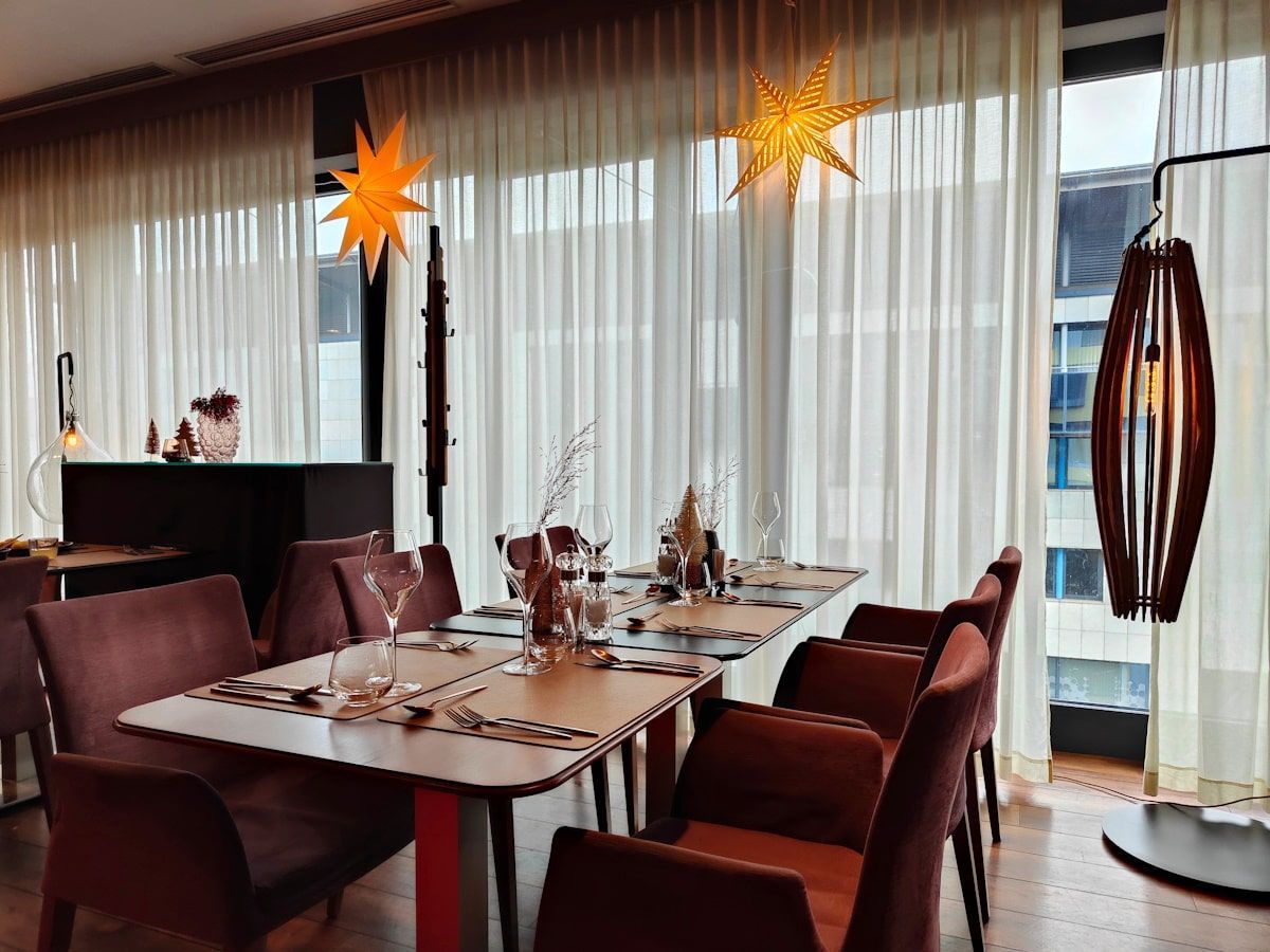 Restaurant interior: Dining table with place settings, chairs, and window with curtains. Star-shaped lights hang.