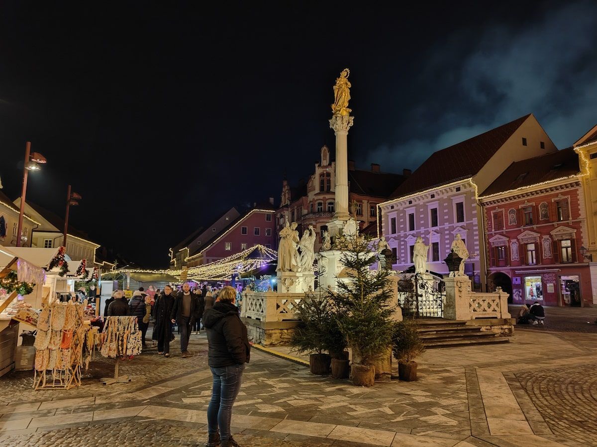 Maribor Christmas Night scene: Festive square with Christmas decorations, people, and a decorated monument.