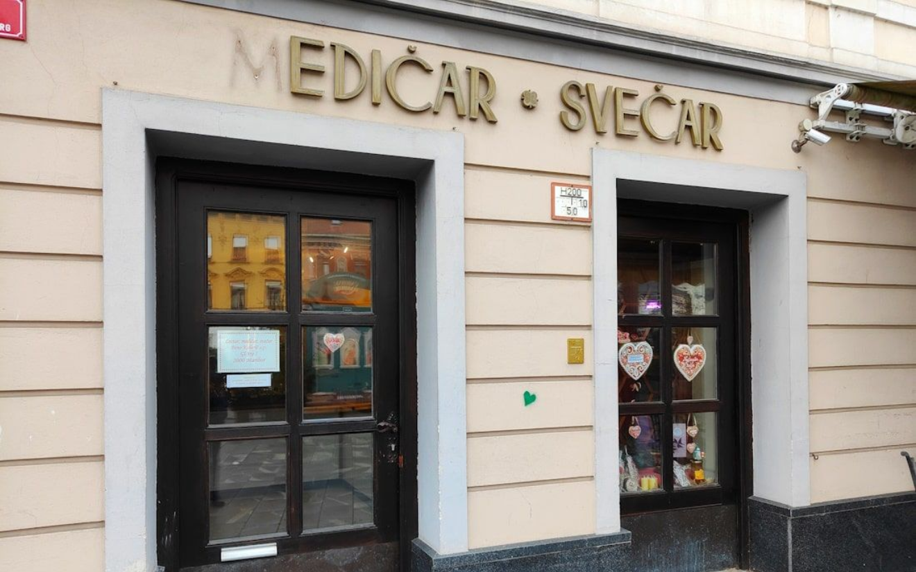 Maribor Exterior of a shop with a dark door, sign reads “MEDICAR • SVEČAR”. Beige building with gray trim and cobblestone.