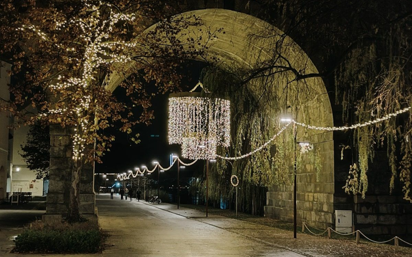 Maribor Night view of a walkway under an arch, decorated with string lights. A tree on the left also has lights.
