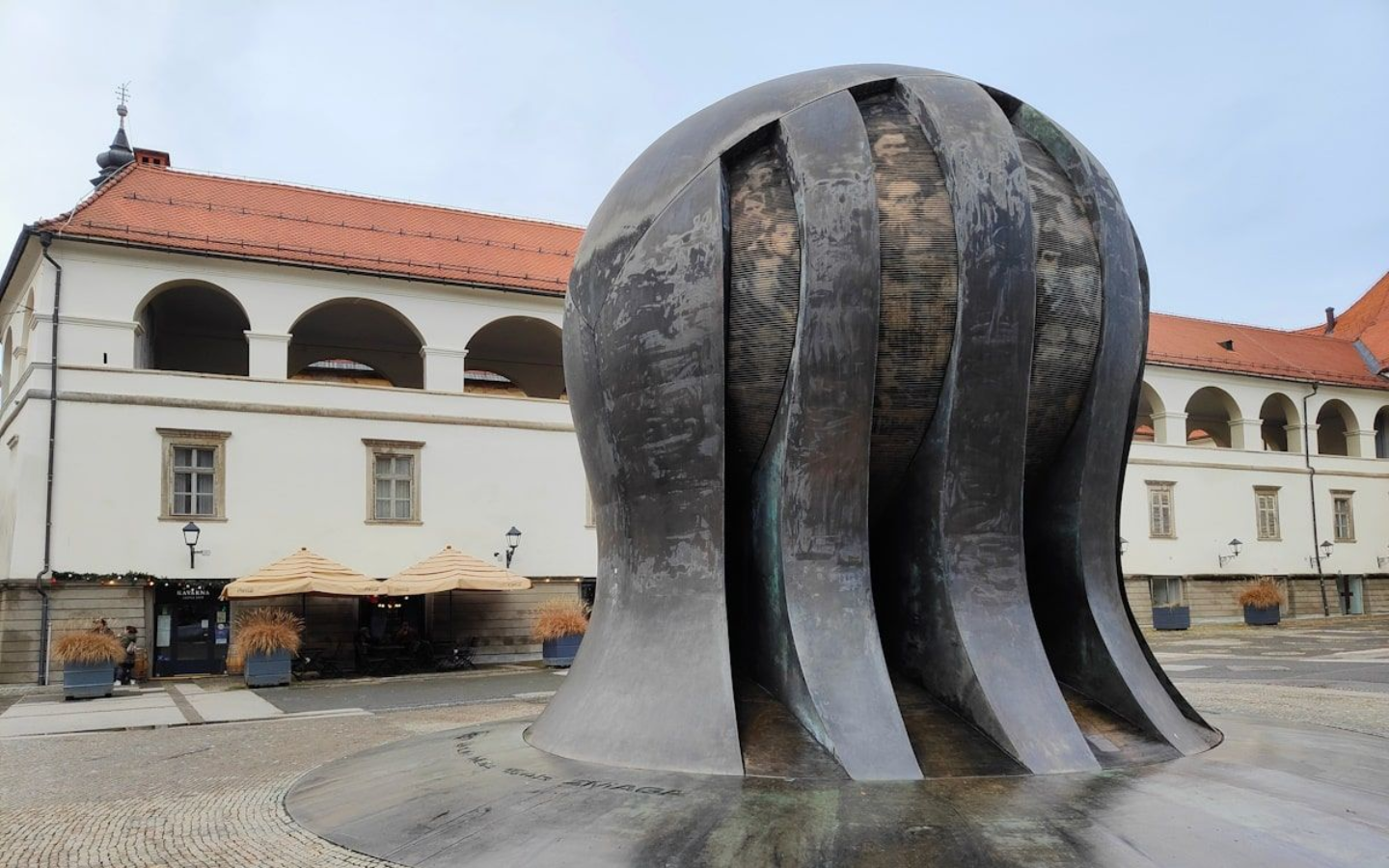Maribor Denkmal Bronze sculpture with vertical slits, in front of a white building with red tile roof, on a cobblestone plaza.