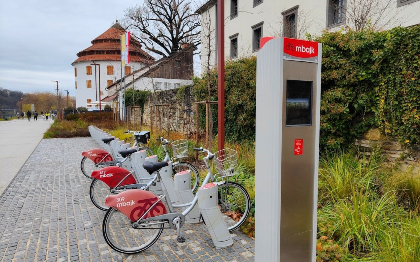 Maribor Fahrräder Bikes in a rental station along a paved walkway; old building with red accents in the background.