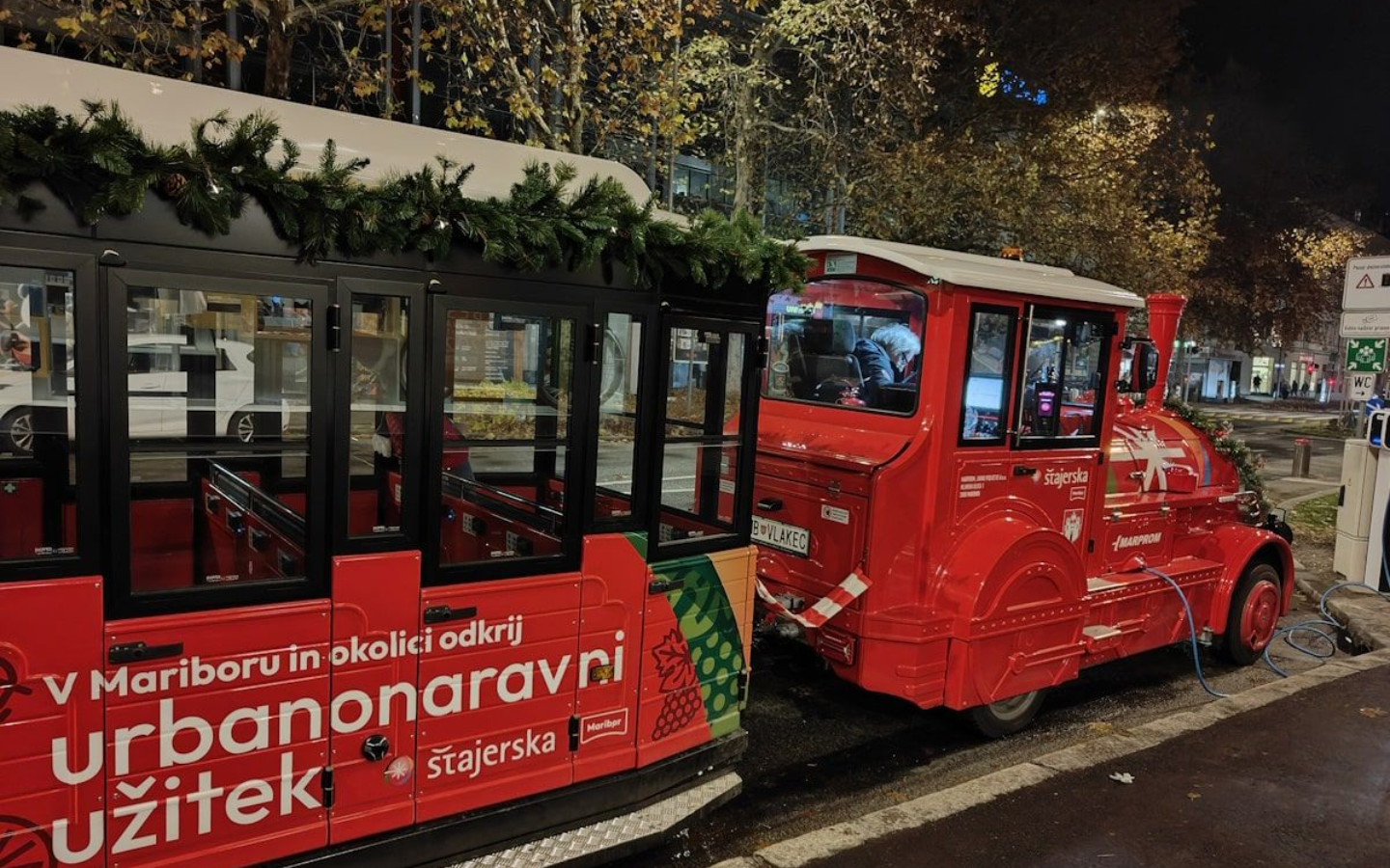 Eletro-Bummelzug Red Christmas train decorated with garland; parked on a city street at night.