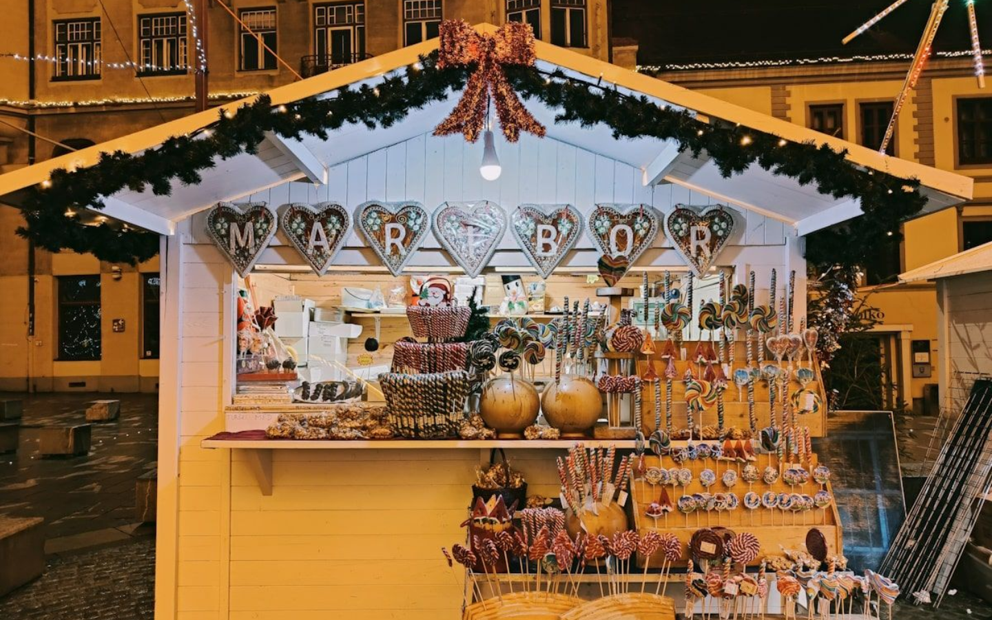 Weihnachtsmarkt Maribor Night scene of a Christmas market with string lights, people, tables, and buildings.
