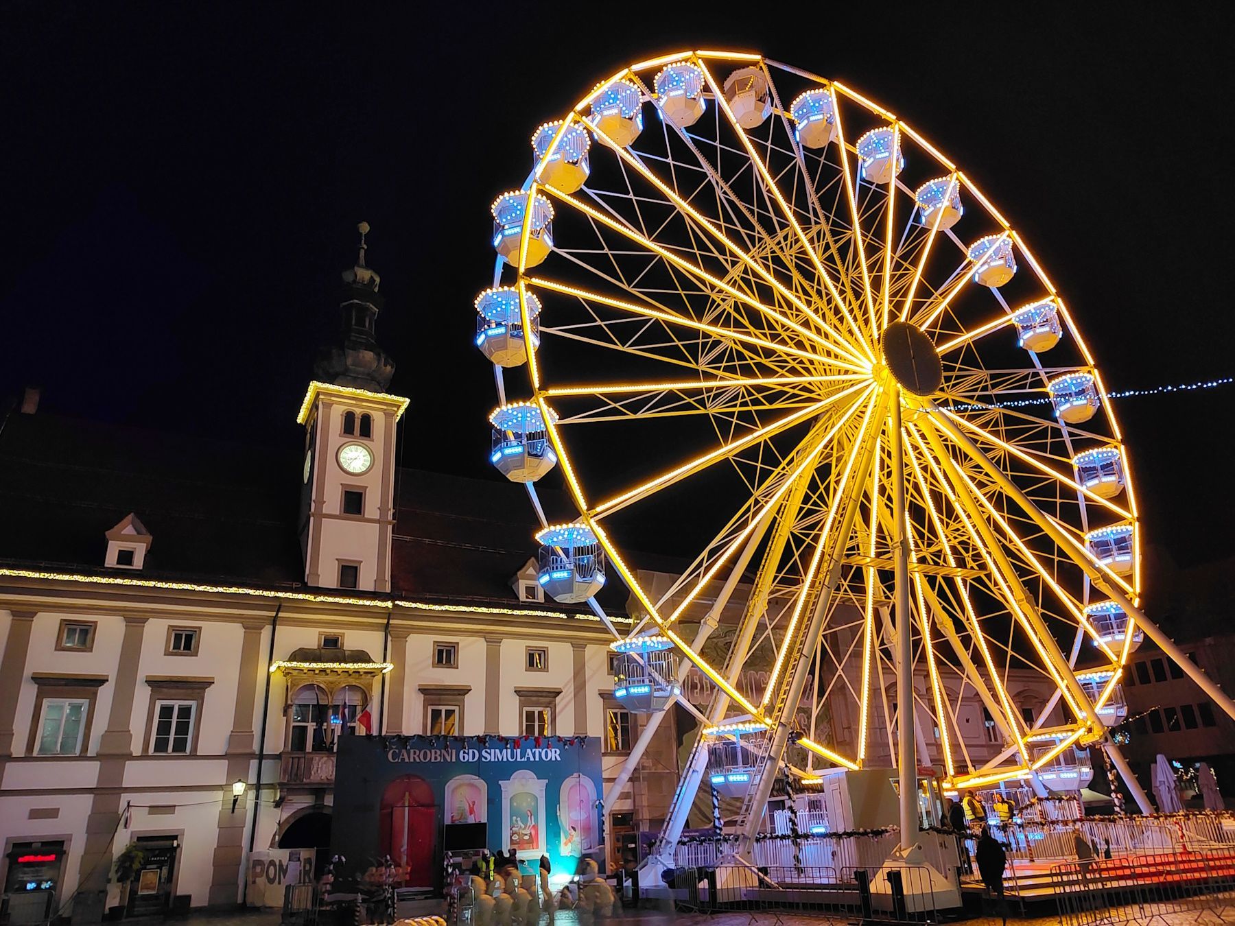Ein mit gelben und blauen Lichtern beleuchtetes Riesenrad steht nachts vor einem Gebäude.