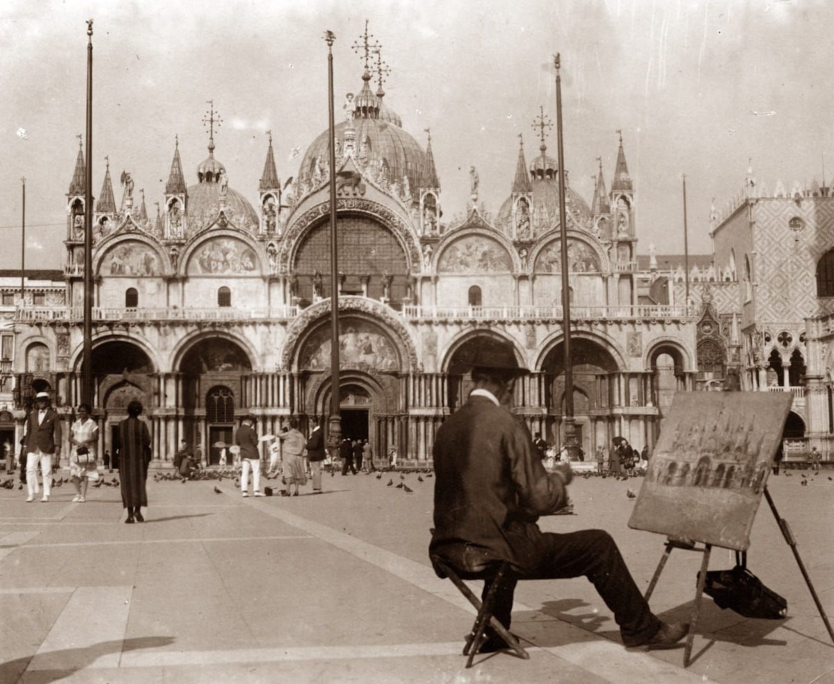 A man sits on a stool sketching St. Mark's Basilica in Venice on a paved square with other people and pigeons nearby.
