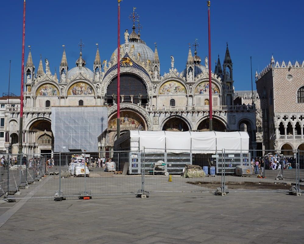 St. Mark's Basilica in Venice, with construction scaffolding and white barriers in front of the ornate facade under blue sky.
